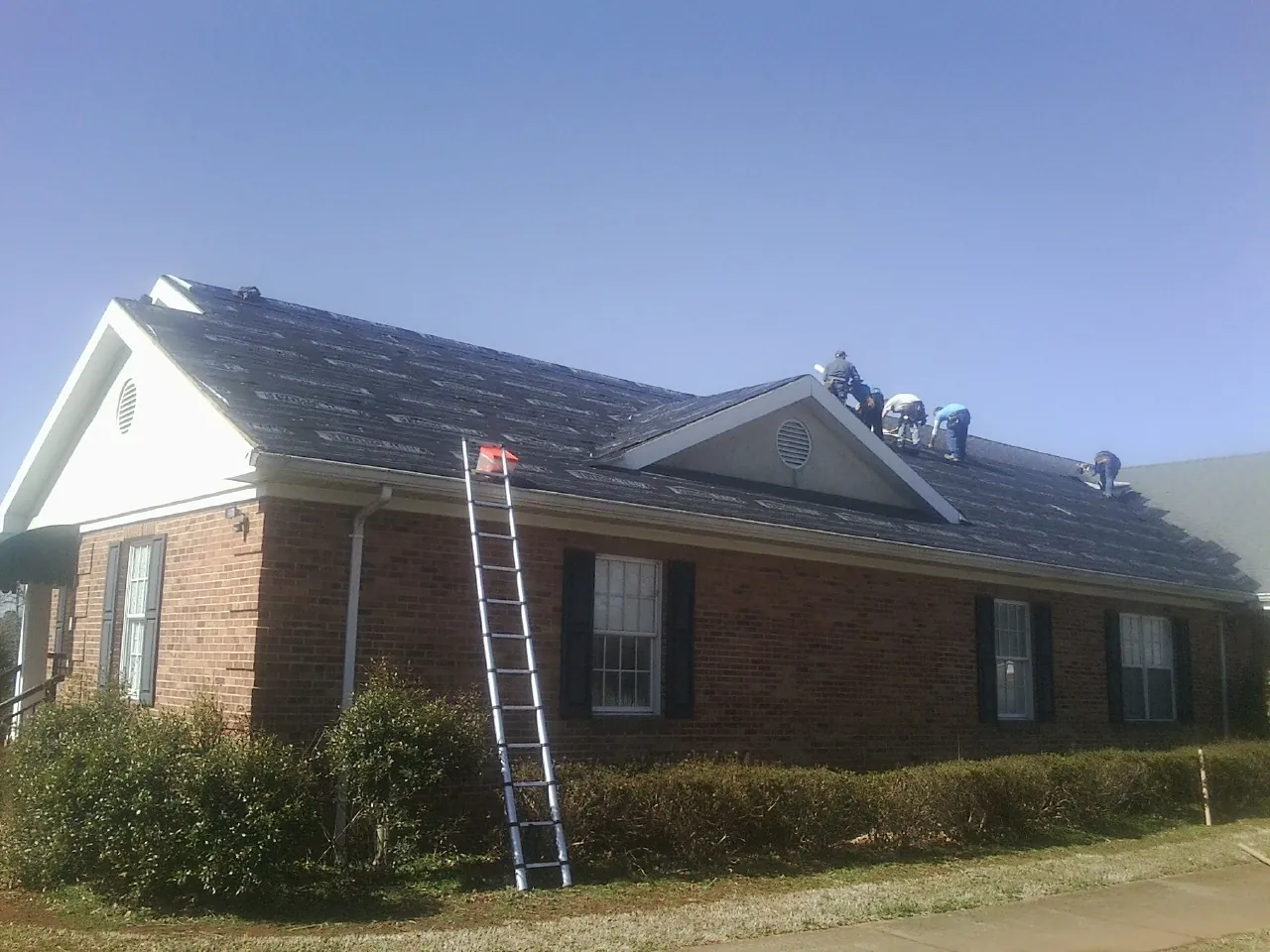 Crew tearing shingles off a brick ranch roof with a ladder set — Emergency Roof Repair work by our crew in Ten Mile