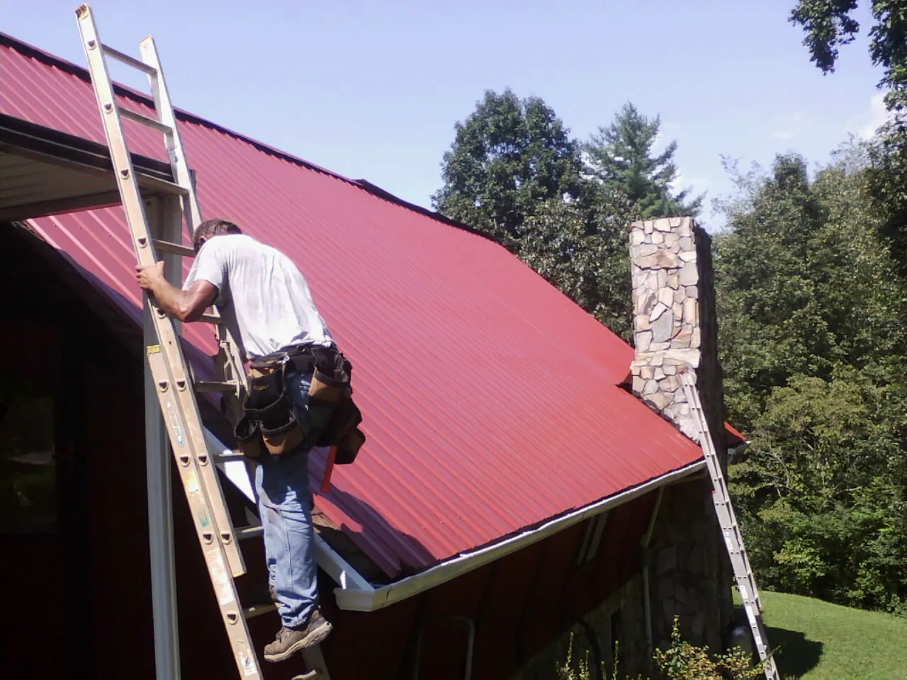 Roofer on a ladder beside a red metal roof and stone chimney — Soffit & Fascia Repair by Roofing Brothers in Ten Mile